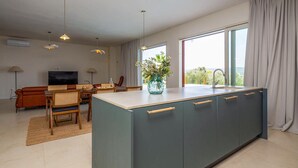 A modern kitchen with a large island, a dining area with wicker furniture, and a view of greenery through large windows. - Cozy home in Otok Babac with private swimming pool, can be inside or outside (Otok Babac)