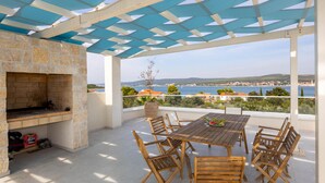 An outdoor patio with a stone fireplace, wooden table and chairs, and a view of the sea and town. - Cozy home in Otok Babac with private swimming pool, can be inside or outside (Otok Babac)