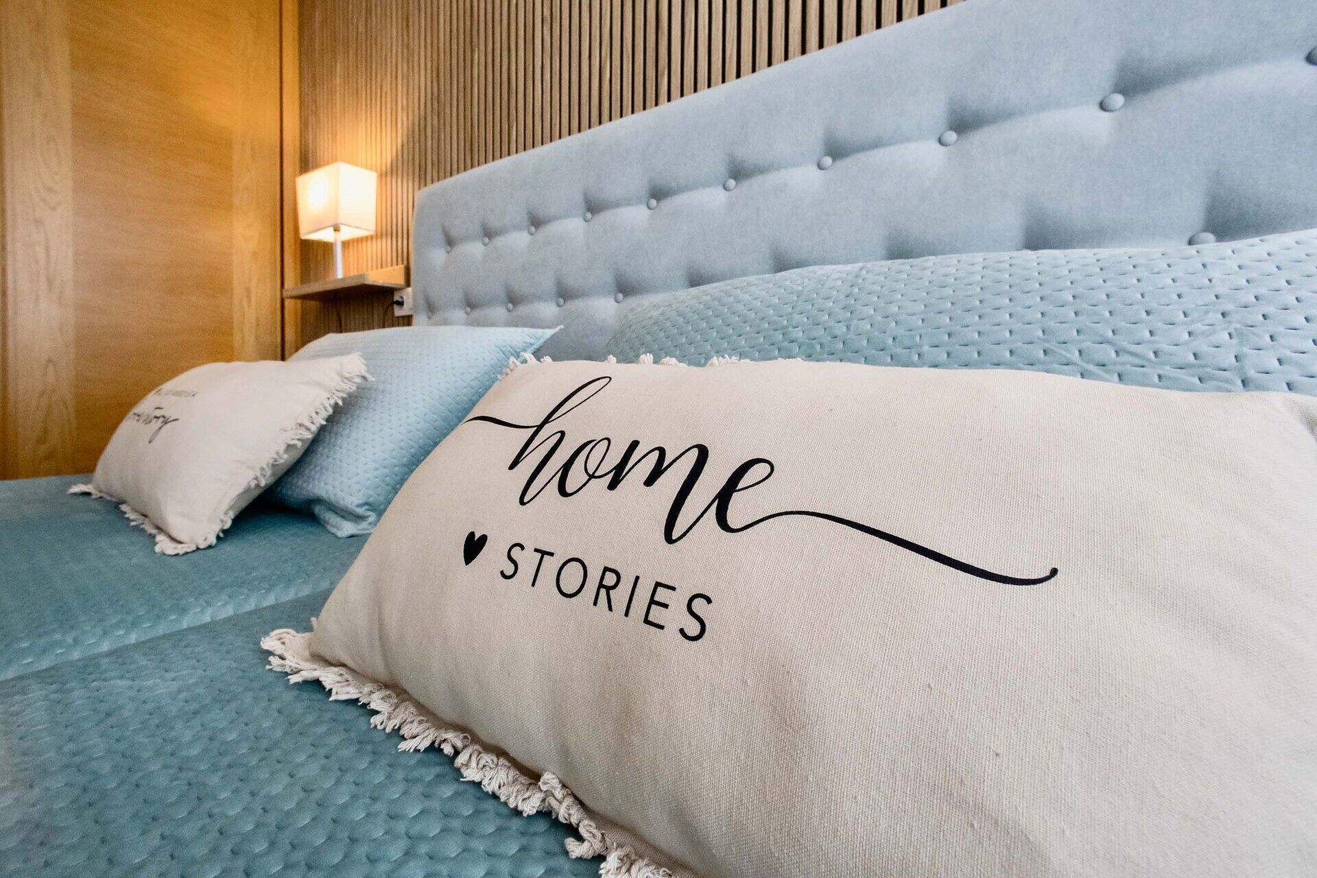 Una cama con cabecera capitonada, una colcha acolchada en tono azul claro y una almohada color beige con la inscripción 'home STORIES'.