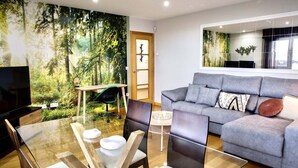 Un salon moderne avec une fresque représentant une forêt, un canapé gris, une table à manger en verre et un téléviseur.