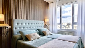 Une chambre à coucher avec une tête de lit capitonnée, deux lits, une vue sur la ville grâce à de grandes fenêtres et un mur avec des lambris en bois.