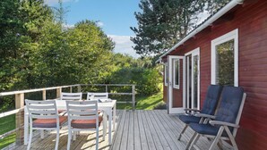 Une terrasse en bois avec une table et des chaises, un bâtiment rouge doté d’une porte coulissante, et une vue sur les arbres et la verdure.