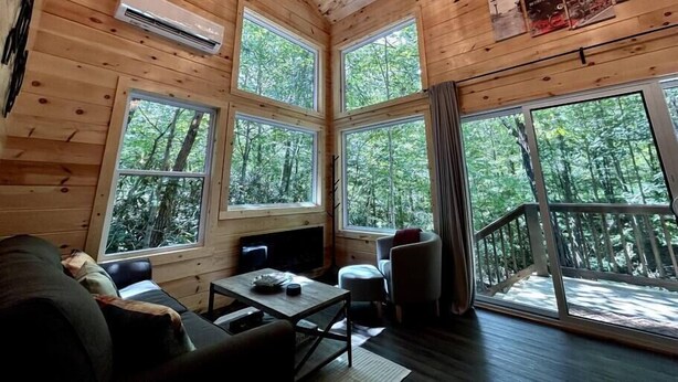 A cabin with wooden walls and large windows, a ceiling fan, a TV, and a dining area. - Boxcar Cabin - Hot Tub, Fire Pit, Train-Theme Stay (Stanton)
