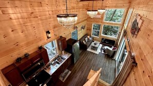 A wooden cabin interior with a kitchen, dining area, and living room, featuring large windows and a chandelier. - Boxcar Cabin - Hot Tub, Fire Pit, Train-Theme Stay (Stanton)