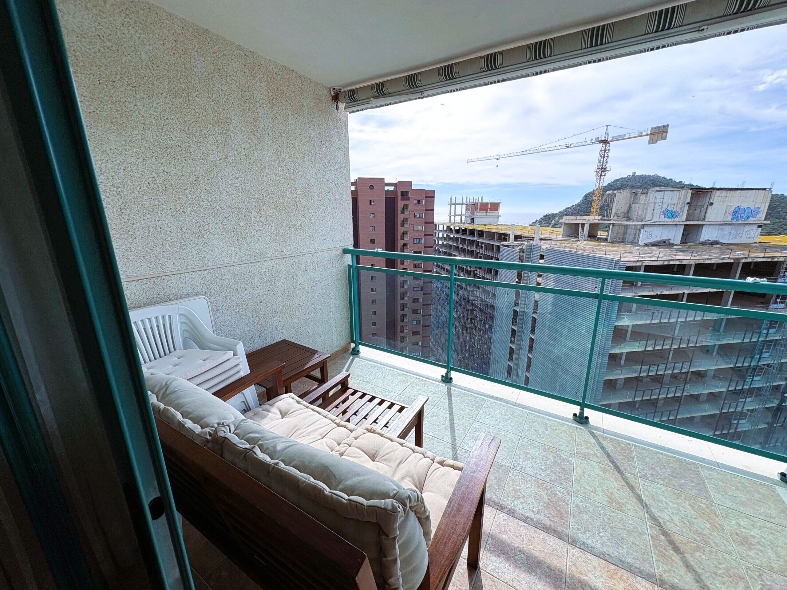 A balcony with a cushioned sofa, a small table, and a view of a construction site with buildings and a crane.