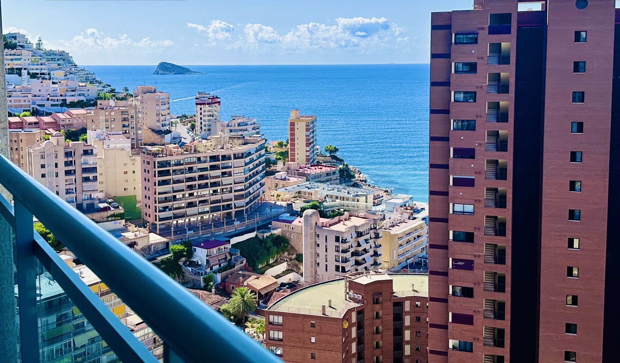 Un paisaje urbano costero con una combinación de edificios residenciales y comerciales, un cielo azul despejado y un cartel que indica 'GARAGE INCLUIDO EN EL PRECI'.