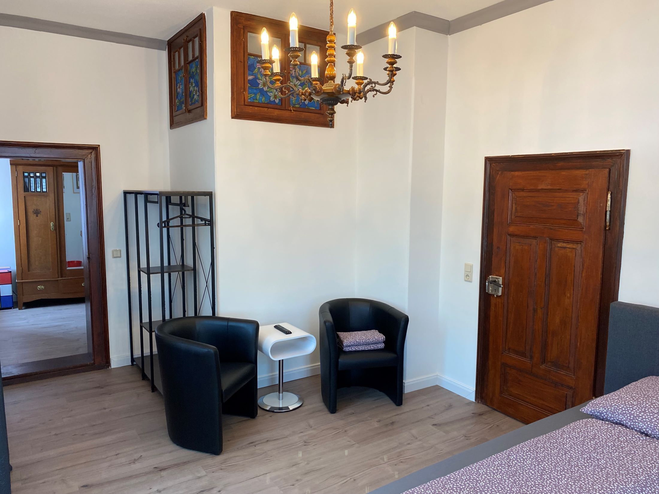 Een kamer met een houten garderobe, een zwarte stoel, een klein wit tafeltje en een blauw beddegoed met dessin.