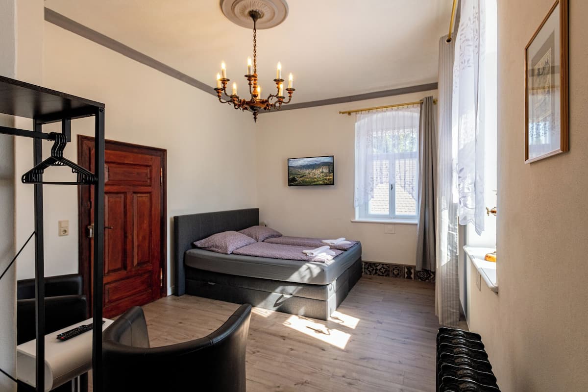 A bedroom with a bed, a desk, a chair, a hanging light fixture, a window with curtains, and a door.