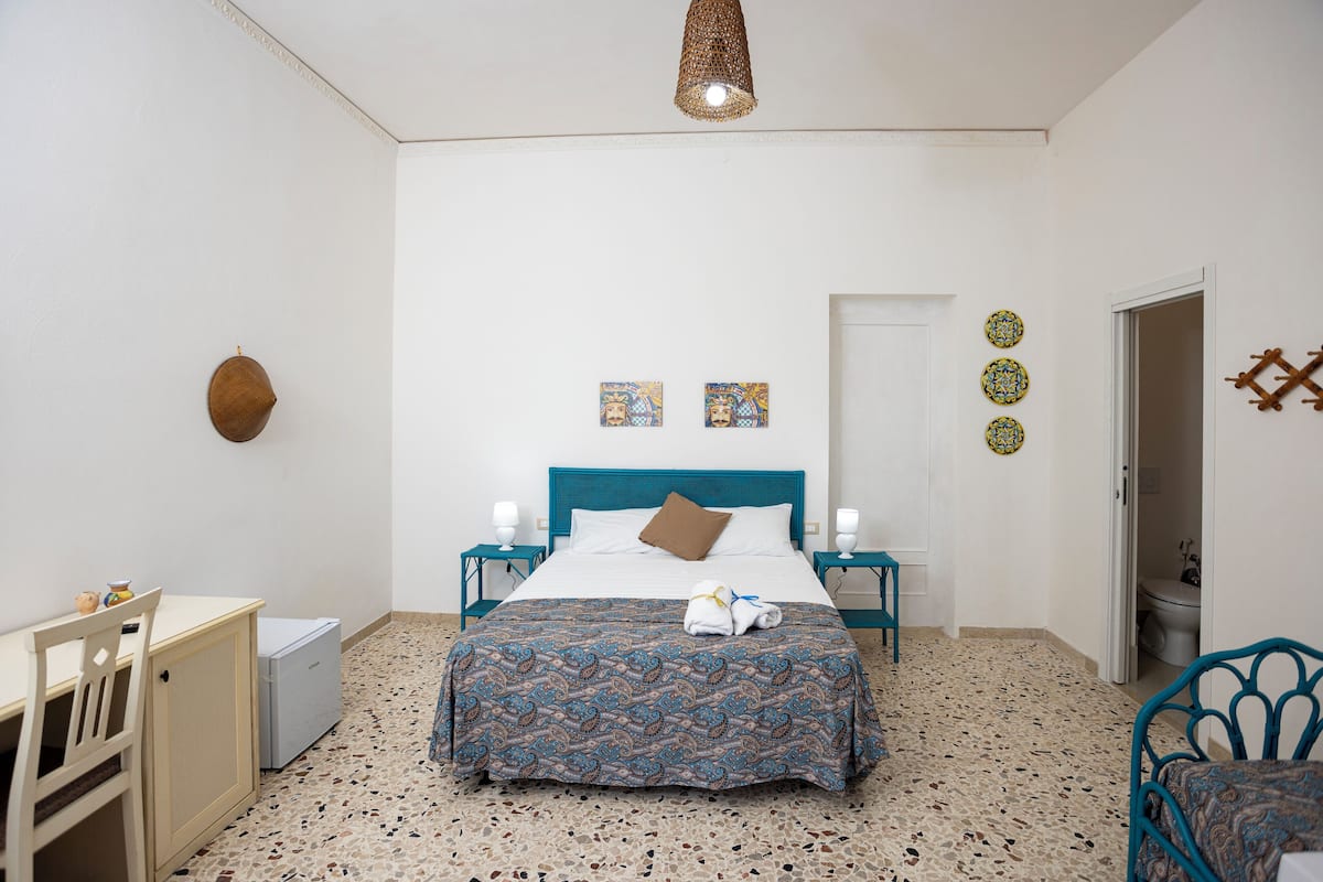 Un dormitorio con cama, dos mesitas de noche, un escritorio, una silla, un armario y una puerta que da al baño.