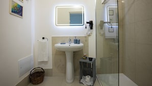 A bathroom with a pedestal sink, a mirror with LED lighting, a glass shower enclosure, and a wicker basket. - SALUTAMU (Misiliscemi)