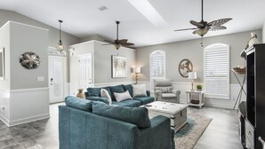 A spacious living room with a teal sofa, a coffee table, and a fireplace. There are two ceiling fans, a large window with blinds, and a decorative wall piece. - Remodeled home minutes to the beach! (Miramar Beach)