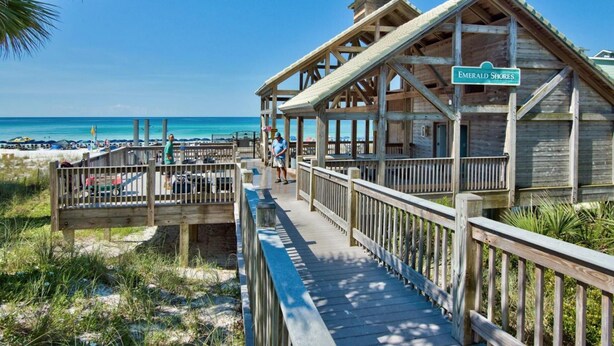 A wooden boardwalk leading to a beach house named 'Emerald Shores' with a view of the ocean and beach umbrellas in the background. - Remodeled home minutes to the beach! (Miramar Beach)