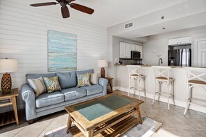 A living room with a sofa, coffee table, and a kitchen area with bar stools and appliances.