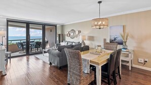 A spacious living area with a dining table, wicker chairs, a grey sofa, and a large window overlooking the ocean. - Updated beach condo with stunning views! (Miramar Beach)