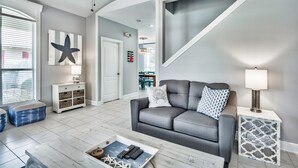 Uma sala de estar moderna com um sofá cinzento, uma mesa de centro e uma mesa lateral com uma luminária. Há uma grande decoração em forma de estrela na parede e uma porta que dá para outro quarto.