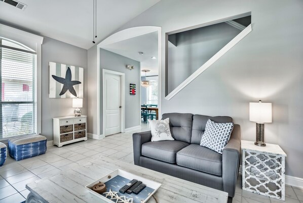 Uma sala de estar moderna com um sofá cinzento, uma mesa de centro e uma mesa lateral com uma luminária. Há uma grande decoração em forma de estrela na parede e uma porta que dá para outro quarto.