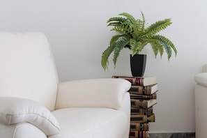 A white armchair, a black vase with a green plant, and a stack of books against a white wall.