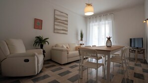 Ein Wohnzimmer mit einer Couch, einem Esstisch mit Stühlen, einem Fernseher und einer Pflanze.