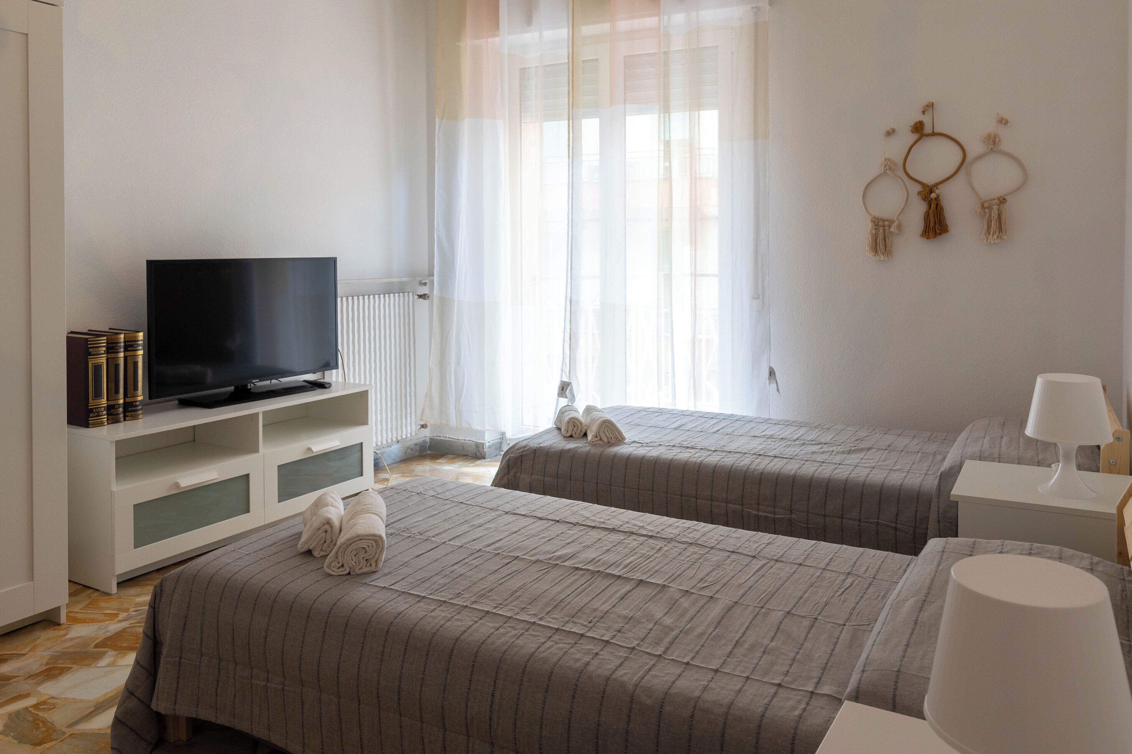 Ein Hotelzimmer mit zwei Betten, einem Fernseher auf einem weißen Schrank, einem Fenster mit durchsichtigen Vorhängen und Wanddekorationen.