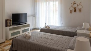 Ein Hotelzimmer mit zwei Betten, einem Fernseher auf einem weißen Schrank, einem Fenster mit durchsichtigen Vorhängen und Wanddekorationen.