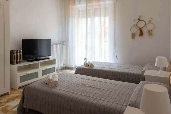 A hotel room with two beds, a TV on a white cabinet, a window with sheer curtains, and wall decorations.