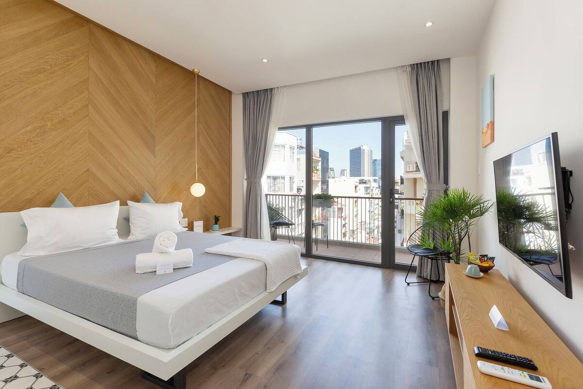 A modern hotel room with a large bed, a TV, a balcony with a city view, and a wooden floor.