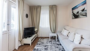 A living room with a white sofa, a TV on a stand, a small side table, and a painting of boats on the wall. - Ben Yehuda By theLight apartment (Tel Aviv)