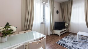 A modern living room with a glass dining table, white chairs, a flat-screen TV, and a small white side table. - Ben Yehuda By theLight apartment (Tel Aviv)