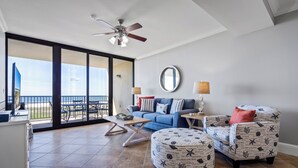 A spacious living area with a large sliding glass door leading to a balcony, a ceiling fan, a sofa, a chair, a coffee table, and a side table with a lamp.