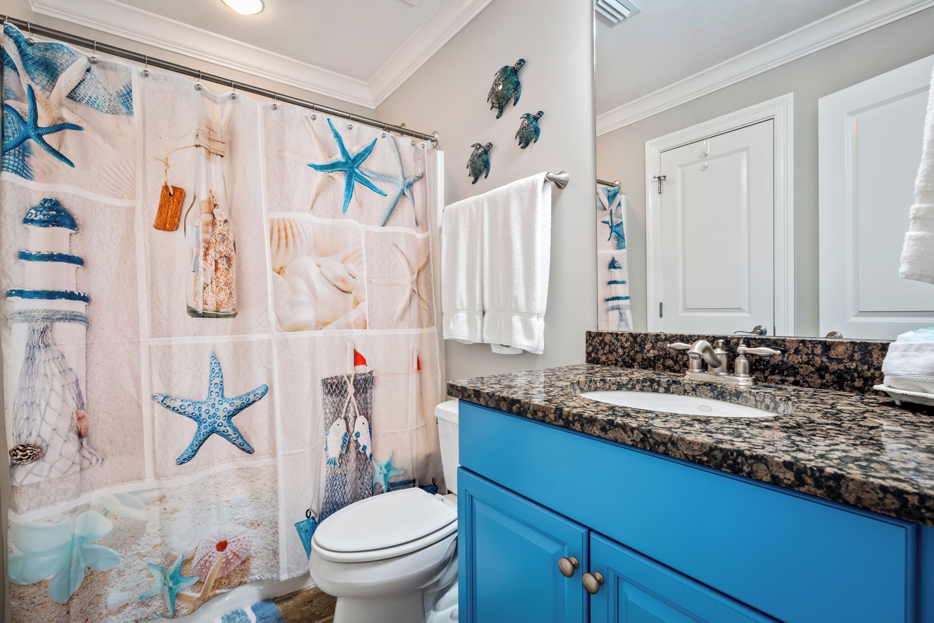 A bathroom with a shower curtain featuring sea-themed designs, a white toilet, blue cabinets, a granite countertop, and a mirror above the sink.