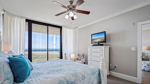 A bedroom with a bed, a television, a ceiling fan, and a view of the outdoors.