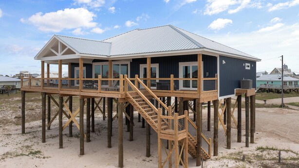 Een houten huis met een terras en metalen dak, gebouwd op palen boven zandgrond.
