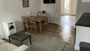 Una sala de estar con una mesa de comedor de madera, sillas, un televisor montado en la pared, un mueble blanco pequeño y un sofá.