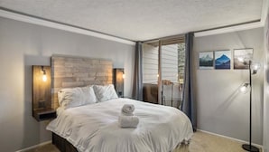 A bedroom with a large bed, a wooden headboard, two bedside lamps, and a view of a balcony with curtains. - Krystal Villa East 303- Nice, Central to Town (Mammoth Lakes)