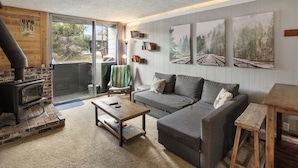 A cozy living room with a fireplace, a gray sofa, a coffee table, and a large window offering a view of the outdoors. - Krystal Villa East 303- Nice, Central to Town (Mammoth Lakes)