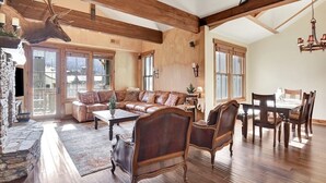 Een ruime woonkamer met een grote bank, een houten eettafel en stoelen. Er is een stenen openhaard en een opgehangen hert.