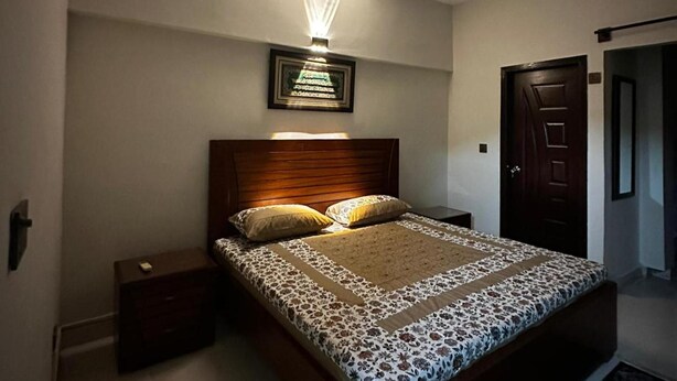 A bedroom with a wooden headboard, a bed with a patterned bedspread, two pillows, a nightstand, a framed picture on the wall, and a door to the right.