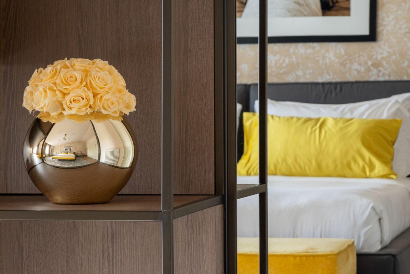 Une pièce avec un lit, un oreiller jaune et un vase rempli de fleurs jaunes, posé sur une étagère.