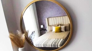 Ein runder Spiegel mit goldener Rahmung an einer Wand, der ein Bett mit gestreifter Bettwäsche, einen Kopfteil, eine Vase mit Trockenpflanzen und ein dunkles Regal darunter reflektiert.