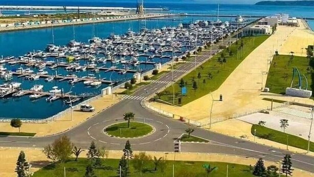 Ein Yachthafen mit zahlreichen dort vertäuten Booten, eine sich um den Hafen schlängelnde Straße und ein Strandabschnitt mit einer Promenade.