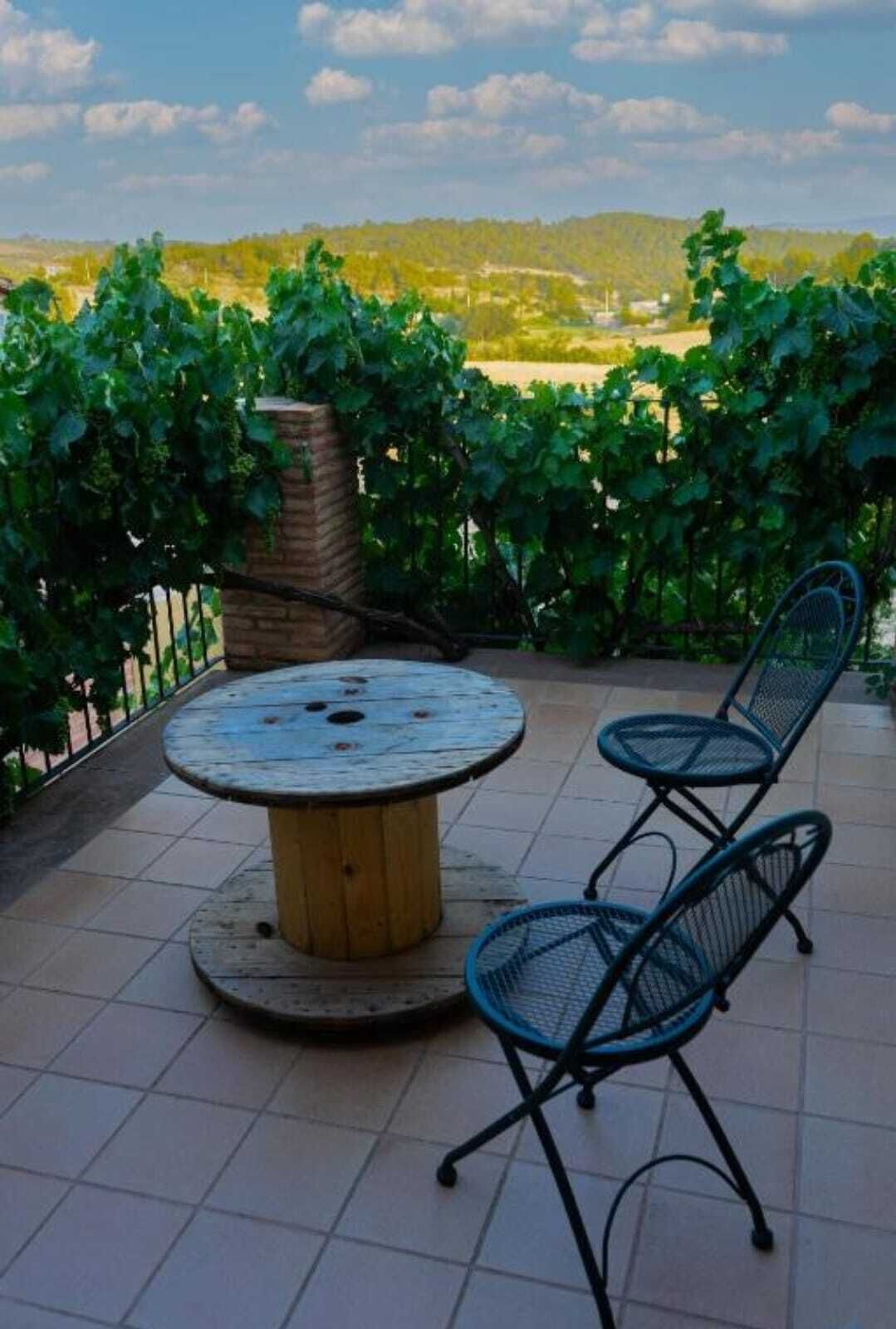Un patio con una mesa redonda de madera y dos sillas azules, rodeado por una jardinera de ladrillo y una valla metálica.