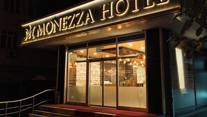 Entrada de um hotel com uma placa destacada que diz "MONEZZA HOTEL" e uma escadaria visível que leva à entrada.