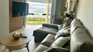 Ett modernt vardagsrum med en soffa, ett litet bord och en tv. Det finns en balkong med utsikt över havet.