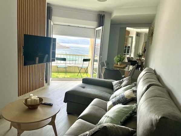 A modern living room with a sofa, a small table, and a TV. There is a balcony with a view of the sea.
