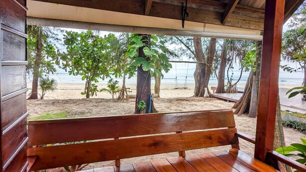 A wooden bench under a covered area with a view of a sandy beach and trees. - Ziggy Stardust (Ranong)