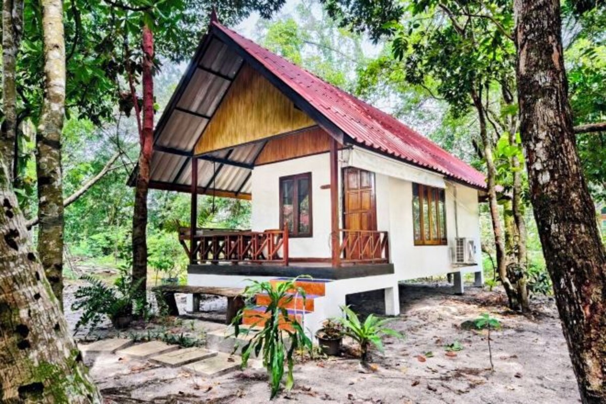 A small, single-story wooden house with a red roof, surrounded by trees and a stone pathway leading to the entrance.