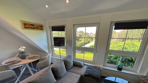 A modern living room with a sofa, a small table, and a chair. There are three windows providing a view of the greenery outside.