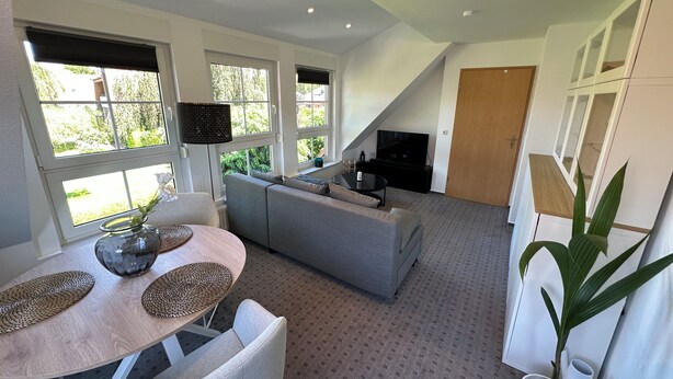 A modern living room with a grey sofa, a dining table with woven placemats, a TV, and a large window with blinds.