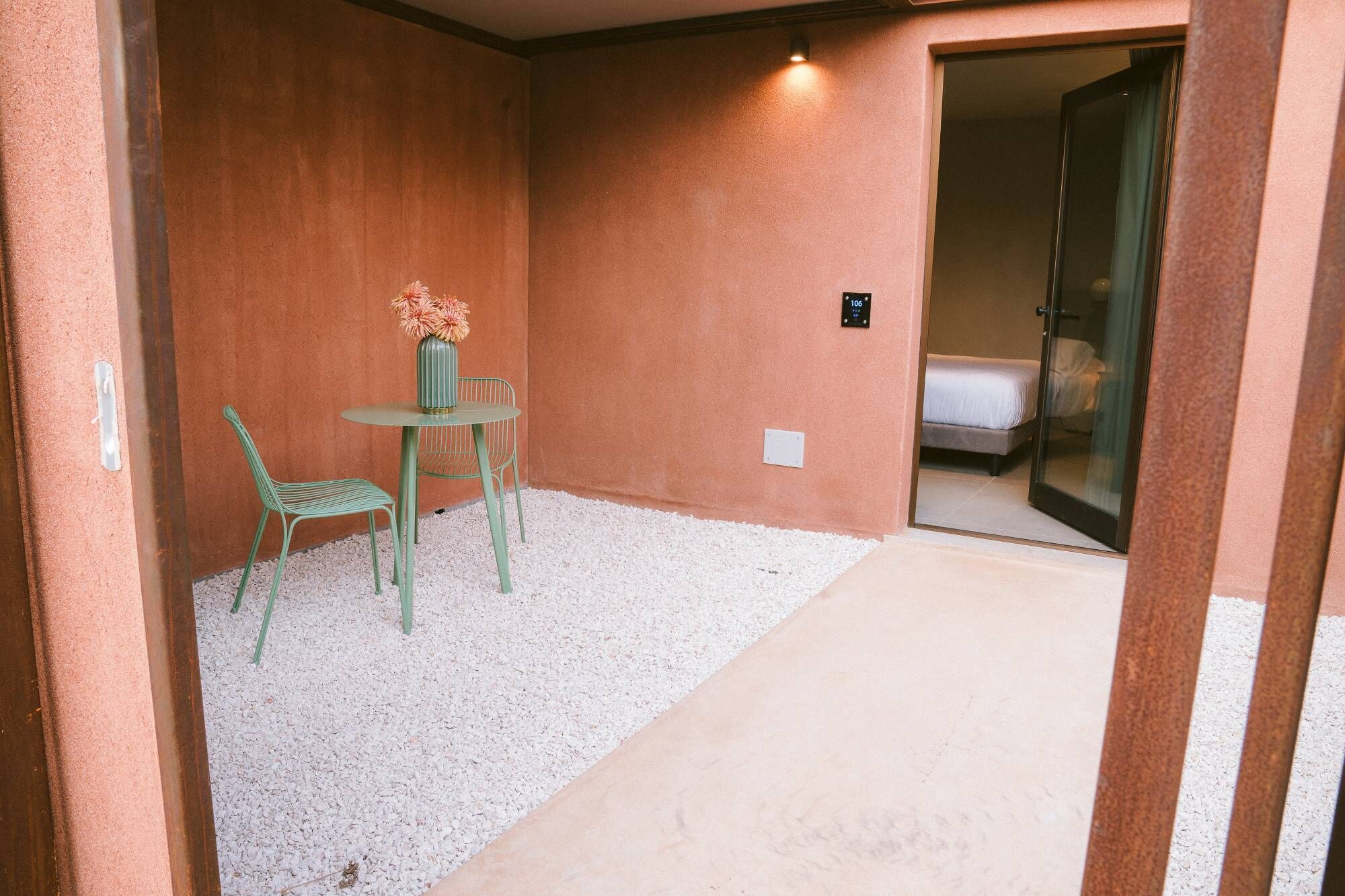 A small, enclosed outdoor area with a table and chairs, a flower vase, and a bed visible through a glass door.