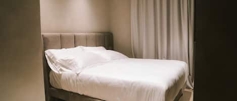 A neatly made bed in a room with a plain wall and a curtain on the right side.